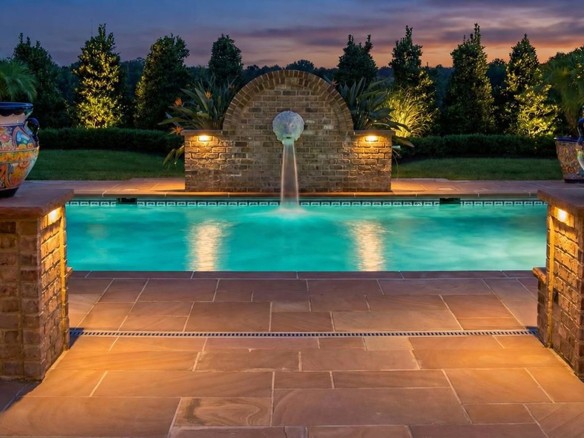 Lit pool with a stone fountain feature and surrounding plantings at dusk